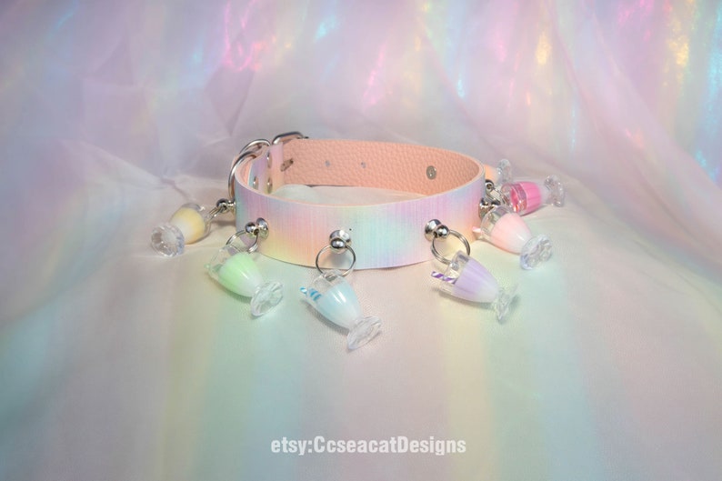 May include: A pastel pink choker with a rainbow tie-dye pattern and silver metal accents. The choker has multiple charms attached, each featuring a clear glass container filled with a different pastel-coloured liquid and a small straw.