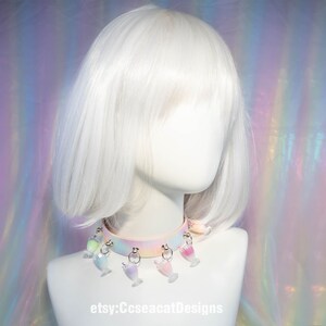 May include: A pastel pink choker with a rainbow ombre effect and multiple charms shaped like colourful glasses. The choker is worn on a white mannequin with a white bob wig. The background is an iridescent, rainbow-coloured fabric.