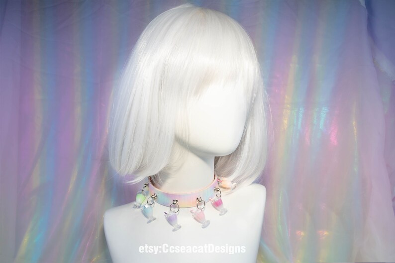 May include: A pastel pink choker with a rainbow ombre effect and multiple charms shaped like colourful glasses. The choker is worn on a white mannequin with a white bob wig. The background is an iridescent, rainbow-coloured fabric.