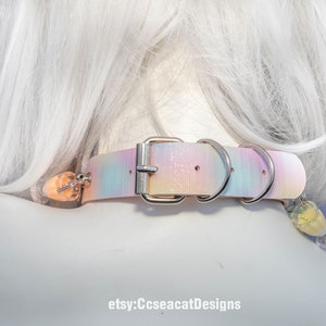 May include: A pastel pink, blue and white iridescent choker with a silver buckle and two silver rings. The choker has a small pink and white charm attached to it.