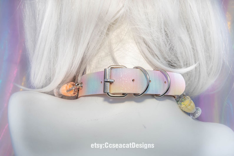 May include: A pastel pink, blue and white iridescent choker with a silver buckle and two silver rings. The choker has a small pink and white charm attached to it.