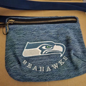 May include: A blue denim crossbody bag featuring an embroidered Seattle Seahawks logo and the text SEAHAWKS in white thread. The bag includes a black zippered top closure with a brass-toned metal ring pull and a long, adjustable dark blue strap.