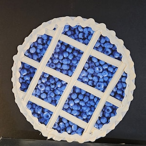 May include: A white fabric pie crust with a scalloped edge, filled with a blue and white blueberry pattern. The crust has a lattice design with white fabric strips.