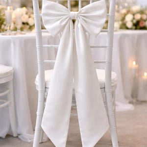Puede incluir: Una silla blanca con un gran lazo elegante atado a la parte trasera. El lazo está hecho de una tela suave, similar al satén, y cuelga en paneles largos y fluidos. La silla es parte de una boda o evento.