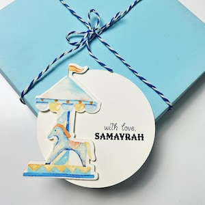 Könnte beinhalten: Hellblaue Geschenkbox mit blau-weißem Garn gebunden. Ein weißer runder Anhänger zeigt eine Karussell-Illustration und den Text "with love, SAMAYRAH". Das Karussell hat ein blaues und gelbes Farbschema.