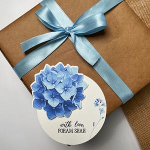 Könnte beinhalten: Ein Geschenk, verpackt in braunes Papier und mit einem hellblauen Satinband gebunden. Ein runder Aufkleber mit einer blauen Hortensienblüte und dem Text "with love, FORAM SHAH" ist angebracht.