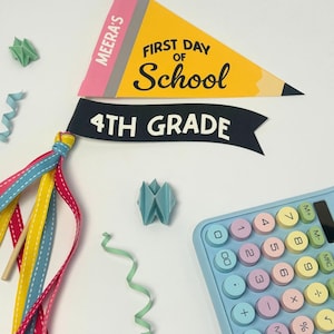 以下が含まれることがあります： MEERA'S FIRST DAY OF SCHOOLと書かれた鉛筆型の黄色いペナント、4TH GRADEと書かれた黒いバナー、パステルカラーのボタンが付いた水色の電卓、木製の棒に結ばれたカラフルなリボン、そして小さな紙の装飾品。