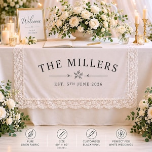 May include: A cream-colored wedding table runner with a lace trim, featuring the text "THE MILLERS" and the date "EST. 5TH JUNE 2026". The runner is 40" x 60" and made of pure linen fabric. A gold framed sign says "Welcome to our forever".