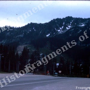 May include: A mountain landscape featuring a dark, forested slope with patches of snow near the peak. In the foreground, a paved road stretches across the frame, bordered by tall evergreen trees and utility poles under a cloudy sky.
