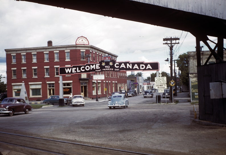 May include: A vintage photo of a red brick building with a sign that reads "Welcome to Canada, Fort Frances Ont."  Cars are driving on the street in front of the building.