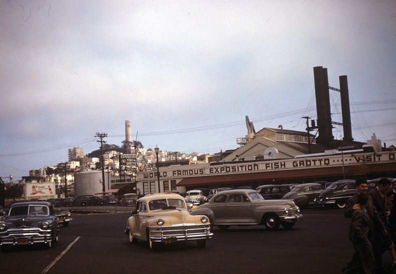 Puede incluir: Un coche vintage de color beige claro estacionado frente a un edificio con un letrero que dice "World Famous Exposition Fish Grotto... Visit Our..." El coche est&aacute; estacionado en una calle con otros coches y gente caminando. El edificio est&aacute; frente a un paisaje urbano con una torre alta en la distancia.