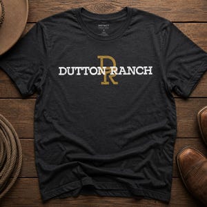 May include: A dark gray t-shirt with the text "DUTTON RANCH" in white and gold lettering. The shirt is displayed on a wooden surface with a cowboy hat, rope, and brown leather boots.