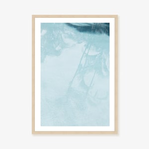 May include: An art print in a light wooden frame, depicting a tranquil scene of palm tree reflections in water. The artwork uses shades of blue, with a light blue base and darker blue details. The overall effect is calming and abstract.