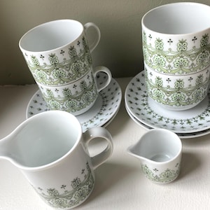 Pode incluir: Um conjunto de chá branco com um design floral verde. O conjunto inclui duas chávenas empilhadas, dois pires, um creme e um pequeno jarro de leite. As chávenas e o creme têm um padrão floral decorativo.