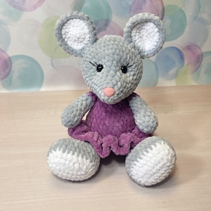 May include: A gray crocheted mouse wearing a purple dress. The mouse has white accents on its ears, paws, and tail.