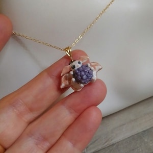 May include: A gold necklace with a pendant depicting a small, fantastical animal clutching a bunch of purple berries. The pendant is set against light pink and white petals. The necklace is shown against a neutral background.