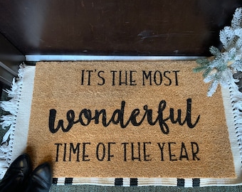 May include: A brown coir doormat with white fringe. The doormat has black text that reads "It's the most wonderful time of the year."