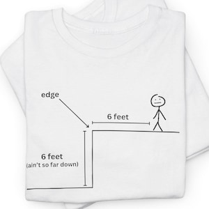 May include: White t-shirt with a black line drawing of a person standing 6 feet away from the edge of a cliff. The text "6 feet (ain't so far down)" is written below the cliff and "6 feet" is written above the cliff.