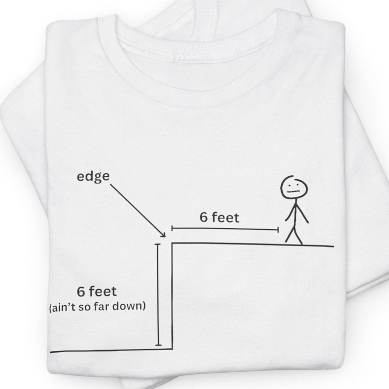 May include: White t-shirt with a black line drawing of a person standing 6 feet away from the edge of a cliff. The text "6 feet (ain't so far down)" is written below the cliff and "6 feet" is written above the cliff.