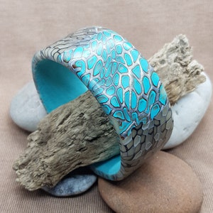 Pode incluir: Um bracelete de cor turquesa e cinza com um padrão e uma superfície texturizada. O bracelete é feito de argila polimérica e é feito à mão.