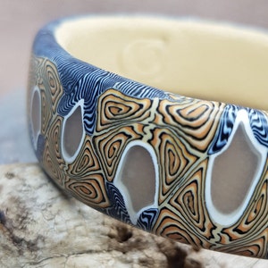 Op de afbeelding: Een kleurrijke armband met een geometrisch patroon in tinten blauw, oranje en wit. De armband is gemaakt van polymer clay en heeft een uniek, abstract ontwerp.