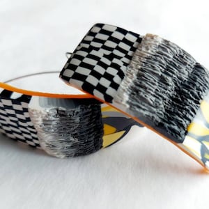 May include: A pair of black and white checkered hoop earrings with a textured gray and yellow design. The earrings are made of polymer clay and have a silver wire hoop.