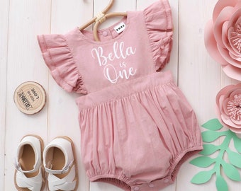 Peut inclure: Une barboteuse rose pour bébé avec des manches à volants et le texte "Bella is One" en blanc. La barboteuse est suspendue à un cintre en bois et se trouve sur un fond en bois blanc.