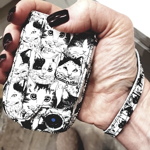 May include: A black and white phone case with a pattern of cats. The case has a blue button on the front.