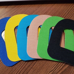 Puede incluir: Siete parches de tela en diferentes colores, incluyendo azul, amarillo, turquesa, beige, verde, negro y verde oscuro. Cada parche tiene una forma rectangular redondeada con un agujero en el centro.