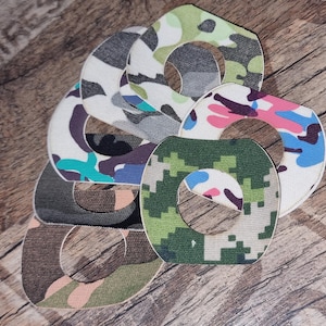 Puede incluir: Una colección de parches circulares de tela con un agujero central, con varios diseños de camuflaje. Los parches muestran colores como verde, marrón, rosa, azul y gris. Los artículos están dispuestos sobre una superficie de madera.