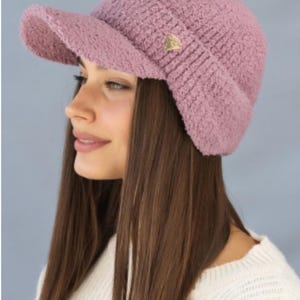 May include: A pink fuzzy knit hat with a visor. The hat has a small gold triangle logo on the front.