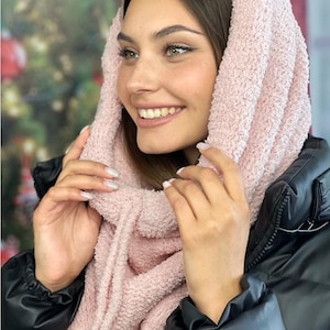 May include: A light pink, textured scarf with an attached hood. The scarf is made of a soft, plush material and is worn around the neck and over the head. The person in the image is wearing a black jacket.
