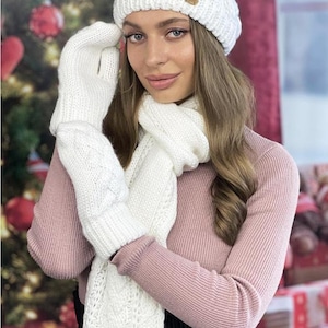 May include: A winter white knit hat, scarf, and gloves set. The hat has a cable knit design with a small wooden tag. The scarf and gloves also feature a cable knit pattern. The set is designed for cold weather.