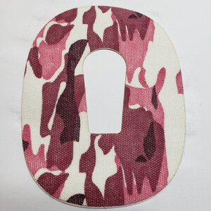 May include: A pink and brown camouflage fabric patch with a rectangular hole in the centre. The patch is shaped like an oval.