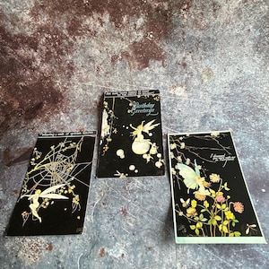 Puede incluir: Tres tarjetas de felicitación vintage con ilustraciones de hadas. Las tarjetas tienen fondos negros con detalles dorados y blancos. Una tarjeta dice "Birthday Greetings". Otra dice "Weaving a web of golden fancies". La tercera tarjeta dice "I dreamed you were with me".