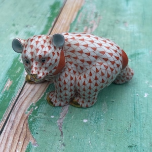 May include: A ceramic bear figurine with a white and orange patterned design. The bear has gray ears, a gold nose, and gold paws. The figurine is on a distressed green and brown wooden surface.