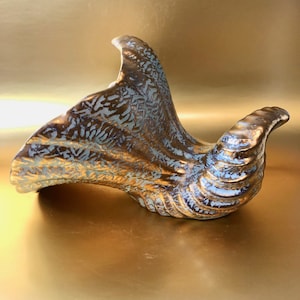 May include: A gold and blue ceramic vase with a textured surface. The vase is shaped like a bird's wing and has a unique, abstract design.