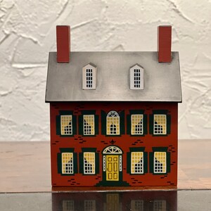 May include: A miniature wooden house painted in red with a gray roof and yellow door. The house has green shutters and white windows. The house is a traditional style with a brick facade.