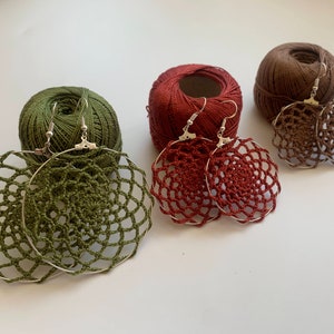 May include: Three pairs of crocheted earrings in green, brown, and red. The earrings are round and have a delicate, lacy design. They are displayed on spools of yarn.