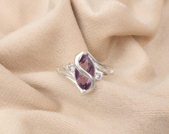 May include: A silver ring featuring a marquise-cut, purple gemstone as the centerpiece. The ring has a swirling design with small, clear accent stones on either side of the main stone. The ring is displayed on a beige, textured fabric.