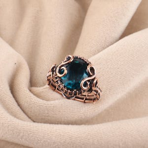 Könnte beinhalten: Ein handgefertigter Kupferdrahtring mit einem großen, runden, teal-farbenen Edelstein. Der Ring hat aufwendige Drahtdetails um den Stein und am Band, vor einem neutralen Stoffhintergrund.