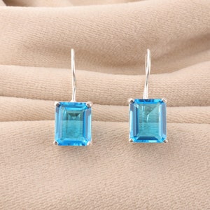 Peut inclure: Une paire de boucles d'oreilles argentées avec des pierres précieuses rectangulaires bleu clair. Les pierres sont serties dans un cadre carré et ont un aspect transparent. Les boucles sont présentées sur un tissu beige.