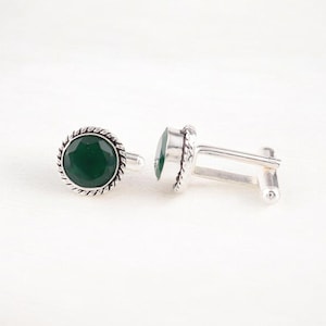 Op de afbeelding: Een paar zilveren manchetknopen met een groene edelsteen in het midden van elke manchetknoop. De manchetknopen hebben een touwachtig ontwerp rond de rand.
