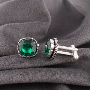 Puede incluir: Un par de gemelos plateados con piedras preciosas verde esmeralda cuadradas. Las piedras están engastadas en un borde decorativo retorcido. Se muestran sobre un fondo de tela gris.
