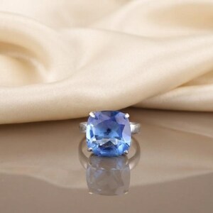 May include: A silver-toned ring featuring a large, square-cut blue gemstone with rounded corners. The stone is held in place by four metal prongs, and the band is a simple, smooth silver-coloured metal that reflects light on the surface below.