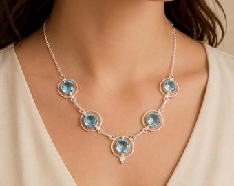 May include: A silver necklace featuring five round, faceted light blue gemstones, each set within a circular silver frame. The stones are connected by thin silver links, creating a symmetrical arrangement that rests along the collarbone.
