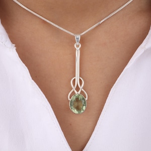 May include: A silver pendant necklace featuring a long, decorative bail with a knotted design and an oval, light green gemstone. The necklace is displayed against a white shirt.