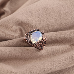 May include: A handcrafted copper wire-wrapped ring featuring an oval, opalescent gemstone. The ring's intricate design showcases detailed wirework around the stone, creating a unique and artistic piece of jewelry. The gemstone has a soft, glowing appearance.