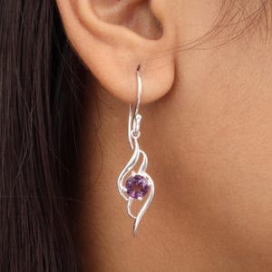 May include: A single silver earring with a purple gemstone. The earring features a curved, flowing design that cradles the round, amethyst-colored stone. The earring is shown worn on an ear.