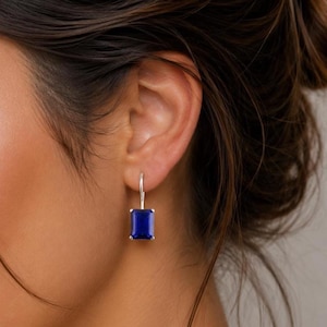 May include: Silver drop earrings with a large, rectangular, deep blue gemstone. The earrings are shown against a background of dark hair and skin, highlighting the vibrant color of the stones.
